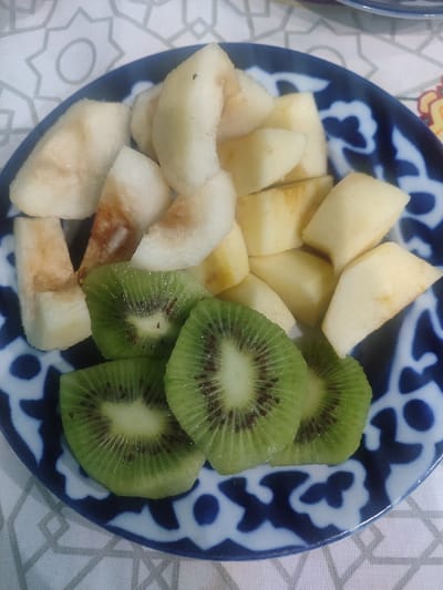 In Rishtan, Uzbekistan, an overhead shot captures a blue and white patterned ceramic plate containing a serving of cut fruit. The plate, featuring a dark blue stylized floral or geometric border pattern on a white background, holds an assortment of approximately 20-25 pieces of light-colored fruit, likely apple or pear, diced into irregular chunks and crescent shapes. Some pieces exhibit slight browning, indicating exposure to air. Arranged at the bottom-left of the plate are four slices of green kiwi fruit, each displaying its characteristic black seeds and pale green core. The plate rests on a light-colored, possibly white, textile surface patterned with repeating grey geometric outlines, suggesting an indoor table setting. No individuals are present in the frame.