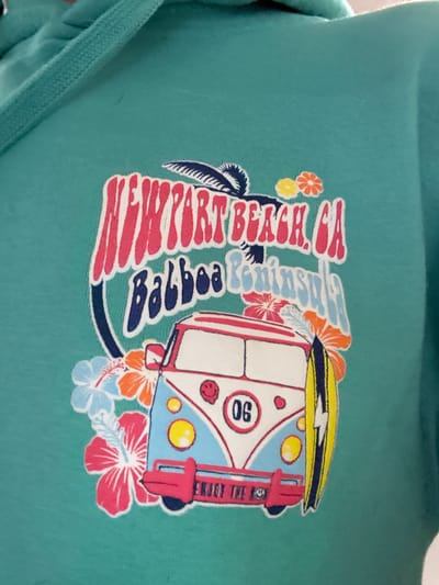 The image displays a close-up of a teal-colored garment, possibly a hoodie or sweatshirt, featuring a vibrant graphic design. The fabric has a soft, textured appearance.
The central subject of the graphic is a stylized vintage surf van, reminiscent of a Volkswagen Microbus, viewed from its front-right corner. The van is predominantly white and light blue, with red outlines and accents, including red taillights and what appears to be a red bumper or frame around the front. A yellow surfboard with a black stripe and white lightning bolt design is strapped to the roof. The front of the van has two round yellow headlights and a circular white emblem containing the number "06" in black. On the rear of the van, partially visible text reads "ENJOY THE RIDE".
Surrounding the van and integrated into the design are elements evoking a tropical, beach-side atmosphere. Large, bold text in red with a white outline reads "NEWPORT BEACH, CA", stacked vertically. Below this, in a light blue script with a white outline, is the word "Balboa". A dark silhouette of a palm tree is visible to the left of the text. Various tropical flowers in shades of orange, pink, yellow, and light blue are scattered around the graphic, adding to the colorful, summer-like feel.
The overall scene depicted by the graphic suggests a relaxed, Californian surf culture, associated with the iconic coastal locations of Newport Beach and Balboa. No specific activity is taking place, but the imagery evokes a sense of leisure, travel, and enjoying the outdoors.