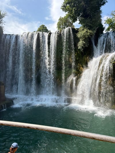 The image shows a majestic waterfall cascading down a rocky cliff face, surrounded by lush green vegetation. The water is a vibrant turquoise color, and mist rises from the point where it crashes into the pool below. A rustic metal railing runs along the right side of the frame, partially obscuring the view of a person wearing a light-colored baseball cap. The sky above is a clear blue with a few scattered white clouds, suggesting fair weather. The scene evokes a sense of natural beauty and tranquility. The orientation of the image, with the waterfall flowing from top to bottom, suggests it was taken from a vantage point near the base of the falls, perhaps from a viewing platform or boat. The presence of the railing and the person indicates that this is a popular tourist destination. The image captures a moment of awe-inspiring natural power and serene beauty.