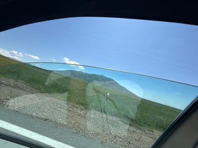 The image shows a view from inside a vehicle, looking out through a slightly open window. The scene outside is a rural landscape with a clear blue sky. In the foreground, there is a dirt and gravel shoulder of a road, with some green grass growing. Beyond that, a gently sloping green hillside stretches into the distance, with a prominent mountain range in the background. The sky is mostly clear, with a few wispy clouds. Power lines are visible stretching across the upper portion of the sky. There are no people or specific structures visible in the image that would indicate a specific location within Artashavan, Armenia. The environment is characterized by rolling hills, vegetation, and open sky.