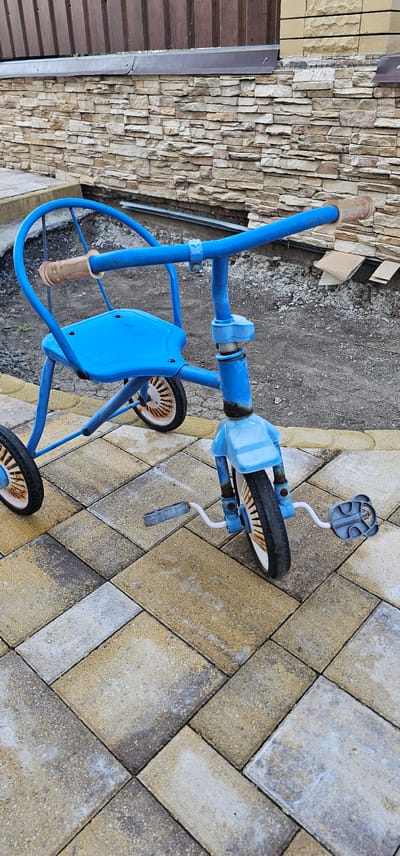 A blue tricycle, showing signs of age and wear with some rust on its white-rimmed wheels, sits on a paved patio made of tan and grey rectangular pavers. The tricycle has a blue seat and handlebars with beige grips. The front wheel has pedals attached. Behind the tricycle, a textured stone wall, possibly part of a building, is visible. To the left, a darker brown slatted surface, possibly a fence or door, can be seen. The ground around the paving stones appears to be dirt. The lighting suggests it is daytime, and the scene is outdoors. The overall impression is one of a well-used children's toy left outside in a yard or patio area in a residential setting.