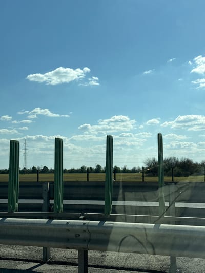 This image, taken near Hegyeshalom, Hungary, captures a bright, sunny daytime scene from the perspective of a moving vehicle on a highway.
The upper part of the frame is dominated by a clear blue sky, dotted with scattered white cumulus clouds. To the right, a grey metal highway guardrail stretches across the image, partially obscured by reflections on what appears to be a car window. These reflections show distorted patterns of the guardrail and possibly interior elements of the vehicle. Several prominent, dark green, vertically-oriented posts, which support the guardrail, are visible in the foreground and mid-ground. The asphalt road surface, with its distinct white lane markings, is visible beneath and behind the guardrail, indicating a highway environment.
Beyond the road and guardrail, a flat, expansive field, appearing golden or light brown, stretches towards the horizon. A line of trees, mostly bare, marks the transition between the field and the sky in the middle distance. Further into the background, a large metal power pylon with its associated transmission lines stands tall against the bright sky.
The strong sunlight casts clear shadows, particularly from the guardrail onto the road, suggesting a time between late morning and early afternoon. The scene conveys a sense of travel through an open, rural landscape under a wide, cloud-speckled sky. There is no visible text in the image.