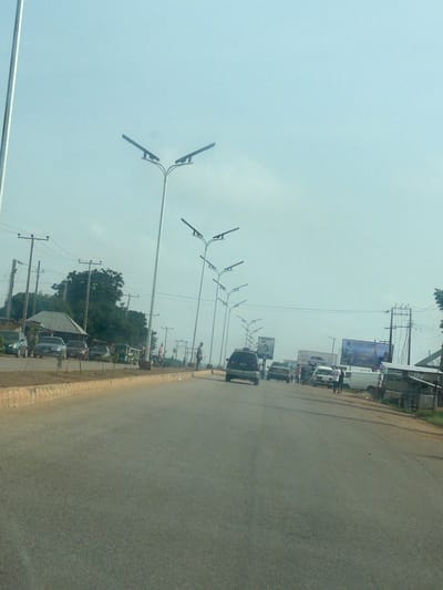 The image shows a street scene in Kassa Market, Nigeria, under a clear sky. The road is paved and lined with a concrete curb and a dirt shoulder. Along the left side of the road, there are several parked cars and what appears to be a small market stall with a corrugated roof. A few trees are visible in the background.

On the right side of the road, there are multiple streetlights with two lamps each, lining the street. Further down the road, there are several billboards, some of which appear to be advertisements. A few people are visible standing on the side of the road, and a few vehicles, including a dark SUV and a white van, are driving or parked on the road. The overall impression is of a bustling market area.