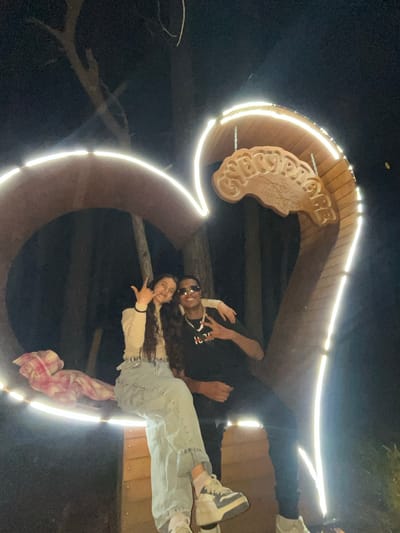 A nighttime shot of a couple posing in front of the entrance sign of Sungurlare, Bulgaria. The entrance is a wooden arch with illuminated LED lights around the frame, with the town's name in a wood-carved sign. The woman is on the right, wearing jeans and making the "rock on" hand gesture. The man is above her, wearing sunglasses and a T-shirt. They appear to be standing inside the arch. The background is dark.