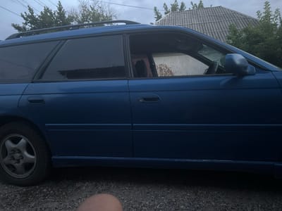 A blue station wagon is parked on a gravel surface. The vehicle's side, from the front wheel well to the rear bumper, is visible. The car has tinted windows, a roof rack, and dark grey side mirrors. The background features greenery, a house with a corrugated roof, and a cloudy sky. The image appears to have been taken in the vicinity of Байсерке, Kazakhstan. A person's knee is visible in the lower right corner of the frame.
