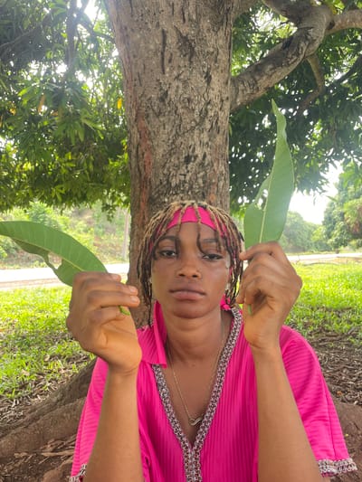 The image, viewed in portrait orientation, depicts a dark-skinned woman in an outdoor, natural setting, looking directly at the camera with a calm expression. She wears her brown dreadlocks pulled back with a bright pink headscarf or bandana, matching her vibrant pink, long-sleeved garment adorned with elaborate silver-colored trim along the neckline and cuffs. A gold-colored necklace with a heart-shaped pendant is visible around her neck. She holds two large, vivid green leaves, one in each hand, seemingly presenting or displaying them; these leaves strongly resemble mango leaves.
The background features a prominent tree trunk with rough, grey bark in the left foreground, which appears to be a mango tree. Its leafy branches extend upwards and across the frame, forming a dense canopy of green foliage through which bright, dappled sunlight filters. This indicates a sunny daytime, likely morning or afternoon. The right side of the background showcases more abundant green vegetation, including grass and undergrowth, suggesting a verdant, tropical environment consistent with the provided location context of Ogbe, Nigeria. There is no visible text in the image.