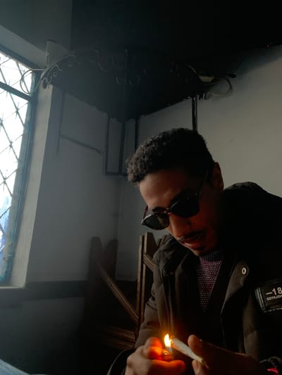 A man in sunglasses, a patterned tie, and a jacket lights a cigarette or joint with a lighter. He is in a room with a window, and there is dark decor and a light-colored wall. The image is dark, likely taken indoors, and tilted.
