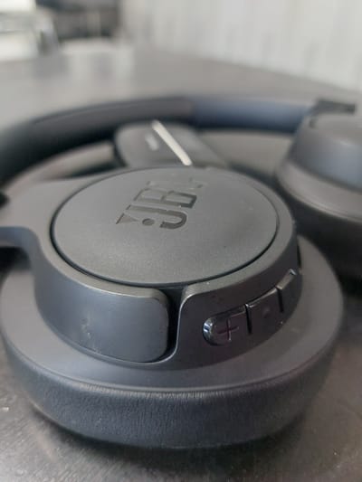 The image shows a close-up, low-angle shot of a black JBL headphone on a dark, reflective surface. The headphone has a textured earcup with the JBL logo prominently displayed in the center. To the right of the logo, there are several control buttons, including what appear to be volume up and down buttons, marked with "+" and "-" symbols. The background is blurred, suggesting a shallow depth of field, and appears to be an indoor environment with some brickwork visible. There is no indication of the city of Алфёрово, Russia, or any people in the image.