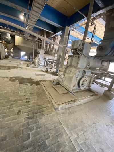The image shows an industrial setting, likely a factory or processing plant, with large machinery and structural elements. The scene is dominated by heavy equipment, pipes, and metal beams, all covered in a layer of dust or fine particulate matter, suggesting an ongoing industrial process. Blue painted structures and metal walkways are visible, indicating the internal architecture of the facility. The lighting suggests it is daytime, likely indoors, as there are no clear indications of natural light sources beyond what might be coming through windows or skylights. The overall impression is one of a functional, perhaps older, industrial environment. There are no people or animals visible in the image. No text is legible. The location context suggests a police facility in Poland, but this information does not directly translate to visual cues within the image itself.