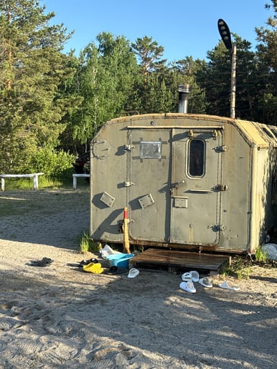The image presents an outdoor scene featuring a robust, olive-green, rectangular structure, resembling a repurposed military or industrial container, possibly functioning as a mobile sauna or bathhouse. It is situated on a wooden platform on sandy, uneven ground. The structure exhibits riveted seams, multiple latches, a small grated window, and a prominent metal chimney extending from its roof. A multi-LED light fixture is mounted on a wooden pole attached to its side.
In the foreground, several pairs of shoes, including white Crocs, yellow Crocs, and dark sandals, are scattered on the ground. One white sandal visibly displays the "Nike" brand logo. A blue bucket and some white fabric, possibly towels, are also near the structure. The background is dominated by dense green pine trees and other conifers, under a clear blue sky. A partial white fence is visible behind the structure.
The scene is brightly lit by natural sunlight, indicating daytime, likely midday or early afternoon, with shadows cast distinctly on the ground. While no individuals are visible, the presence of multiple pairs of shoes, the bucket, and the structure's characteristics suggest an ongoing outdoor recreational activity, possibly a communal bathing or sauna experience in a natural setting. The location context for this scene is Zerendi, Kazakhstan.