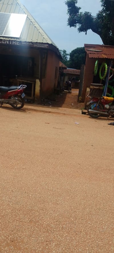 The image depicts a street scene in Baissa, Nigeria. The foreground is dominated by a light brown, pebbled road. In the mid-ground, several buildings are visible. To the left, a building with a corrugated metal roof and a solar panel on its slope has text above its entrance. The text reads: "CENTRE, BAISSA. 08022166012 07033662771." A red motorcycle is parked in front of this building. To the right, another building with a rusty corrugated metal roof displays several green and yellow tires hanging from its side. A red and yellow motorcycle is parked in front of this structure, facing away from the camera, with a wooden crate strapped to its rear. Further in the background, additional buildings and trees are visible, suggesting a semi-rural or town setting. The sky is overcast, indicating a cloudy day. The overall impression is one of a commercial area with small shops and vehicle activity. The scene appears to be during daytime.