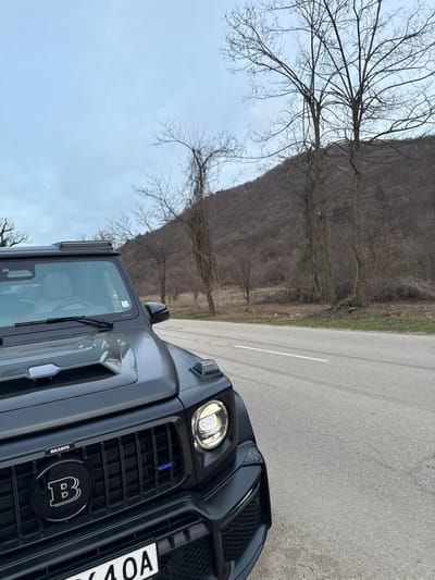 A dark grey Brabus G-Wagen is parked on the paved shoulder of a road in Debnevo, Bulgaria. The vehicle's front-left quarter is prominently featured, displaying a large "B" Brabus logo on its grille and a visible illuminated headlight. A roof-mounted light bar is present above the windshield. The road features a solid white line and tire marks. To the right of the road, a strip of ground leads to several leafless deciduous trees, some heavily covered in vines. A large, dormant tree-covered hill or mountain forms the background under an overcast sky. No individuals or active interactions are visible in the scene.