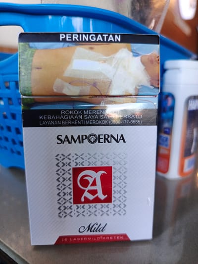 A close-up, eye-level shot shows a pack of Sampoerna A Mild cigarettes positioned vertically in the foreground, resting on a brown textured surface. The cigarette pack is white with a patterned design and features the brand name "SAMPPOERNA" prominently. A stylized red letter "A" is visible within a red square in the center of the pack. Above the brand name, a black band contains text in Indonesian, including "PERINGATAN" (Warning) and information about quitting smoking. The upper portion of the pack displays a graphic image of a person's torso with medical bandages.
Behind the cigarette pack, a bright blue plastic basket is visible on the left, and a white bottle with an orange label (partially obscured) is on the right. The background is blurred and out of focus, suggesting an indoor or shaded outdoor environment. The scene appears to be a still life arrangement, with no people or discernible action taking place. The lighting is adequate, highlighting the details of the cigarette pack. The overall impression is that of a product shot, possibly taken in a market or shop setting. The presence of a blue basket and a white bottle suggests a retail or domestic context, potentially in Bireuenmeunasahdayah, Indonesia, given the product's origin.