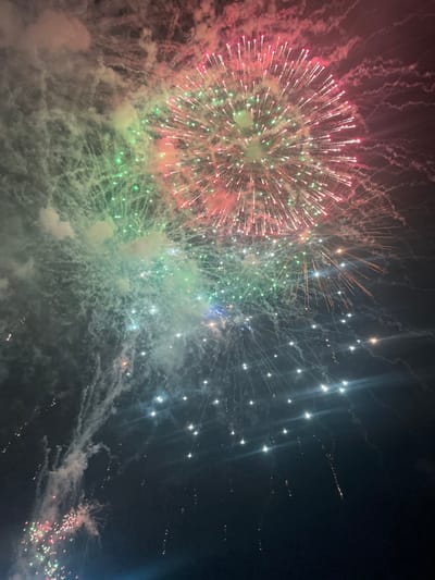 Vivid fireworks illuminate the night sky. A large burst of red, green, and white dominates the left, exhibiting a spherical explosion. To the right, another firework explosion trails off with shining sparks, contrasting against the dark expanse. Smoky clouds intermingle with the light displays, adding texture and depth to the composition. The vibrant colors and dynamic forms create a festive and celebratory atmosphere.