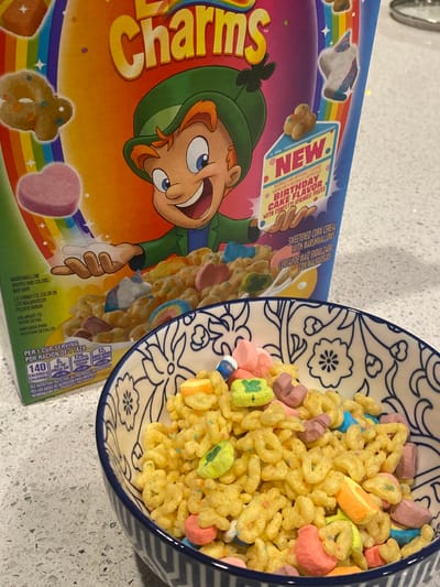 The image displays an indoor scene, likely a residential kitchen, featuring a box of cereal and a bowl of cereal. While the prompt specifies Ridgefield, United States, no discernible geographical markers for this location are present within the image frame.
In the foreground, a ceramic bowl with a blue and white patterned rim is filled with Lucky Charms cereal. The cereal consists of yellow, O-shaped pieces mixed with various multicolored marshmallows, including green shamrocks, pink hearts, blue moons, yellow cake slices with brown bases, and other abstract shapes. Small, colorful confetti sprinkles are visible throughout the cereal.
Behind the bowl, positioned to the left, stands an upright box of "Lucky Charms" brand cereal. The box prominently features "Lucky the Leprechaun" and a rainbow, along with a "NEW Birthday Cake Flavor" label indicating "sweetened corn cereal with marshmallows and confetti sprinkle pieces." Nutritional information and product details are visible on the lower left side of the box.
Both the cereal box and the bowl rest on a light gray countertop surface, which exhibits a speckled or mottled texture consistent with granite or quartz. No individuals are present in the scene, and no specific actions are being performed. The lighting suggests an overhead source, casting minimal shadows.