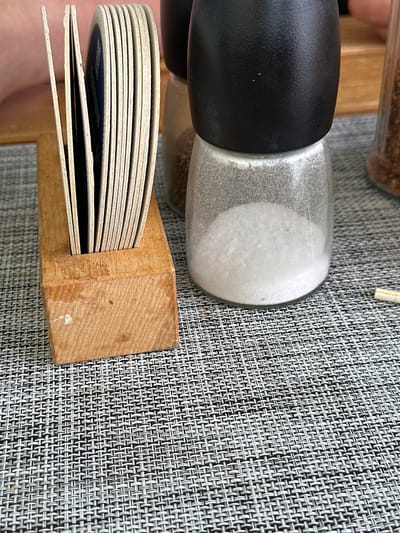 A close-up, eye-level shot shows a collection of coasters in a wooden holder on a textured gray placemat. To the right of the coasters is a glass salt grinder filled with white salt, topped with a black grinder mechanism. Further to the right, a second, less defined glass grinder contains darker particles, possibly pepper. The background shows a blurred wooden table surface. The scene suggests a dining or bar setting in Berchtesgaden, Germany, likely a restaurant or beer garden, given the coasters and grinders.