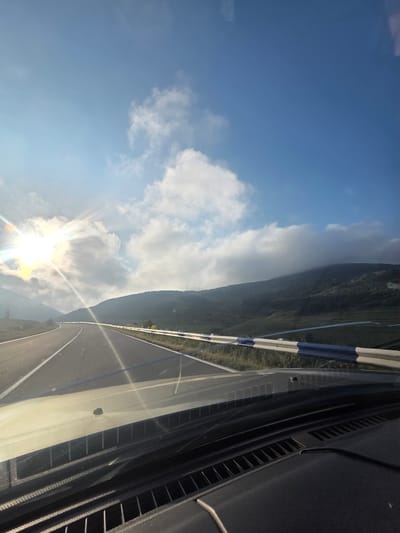 The image depicts a scenic view from a car driving on a road, presumably in or near Jajuravan, Armenia. The sun is bright and appears to be rising or setting, casting a strong glare and sunbeams across the frame. The road curves ahead, bordered by a guardrail with alternating blue and white sections. On the right side of the road, there are rolling hills covered in green vegetation. The sky is a vibrant blue with scattered white clouds. The dashboard of the car is visible in the foreground, suggesting the image was taken by a passenger or the driver. There are no people or animals clearly visible in the scene.