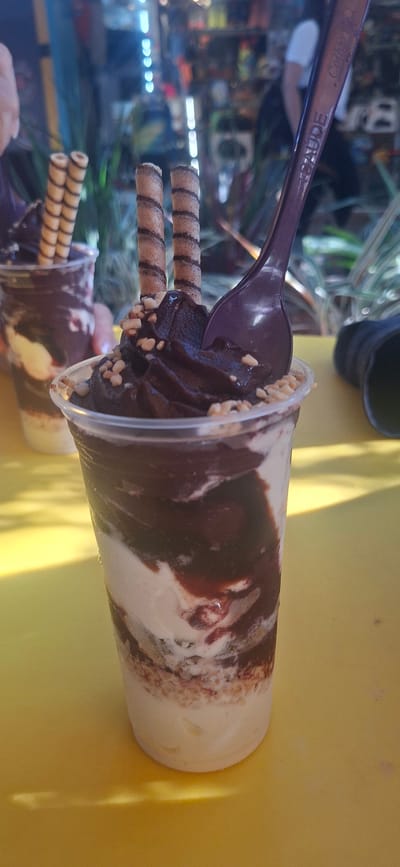 The image captures a close-up of a vibrant dessert served in a clear plastic cup, set against a bright yellow surface, likely a table. The dessert is a swirl of creamy vanilla and rich chocolate soft serve or ice cream, generously covered with a dark chocolate syrup and sprinkled with chopped nuts, possibly peanuts. Two striped wafer rolls are vertically inserted into the dessert, acting as garnishes.
A purple plastic spoon is plunged into the dessert, indicating it is ready to be eaten or is already being consumed. The spoon itself features visible text: "Colibri" and "SAÚDE" (Portuguese for "health" or "cheers"). A second, similar dessert cup is partially visible and blurred in the lower left foreground.
The background is softly out of focus, revealing an outdoor setting with glimpses of green foliage and diffused artificial lights, suggesting a casual eatery, a cafe, or a street vendor in a lively area. The bright lighting implies it is daytime, likely sunny. The entire scene is oriented as if the camera was tilted or the image was rotated, presenting the cup horizontally across the frame. This casual, vibrant presentation of a dessert is characteristic of a warm climate, fitting the context of Armação Dos Búzios, Brazil.
