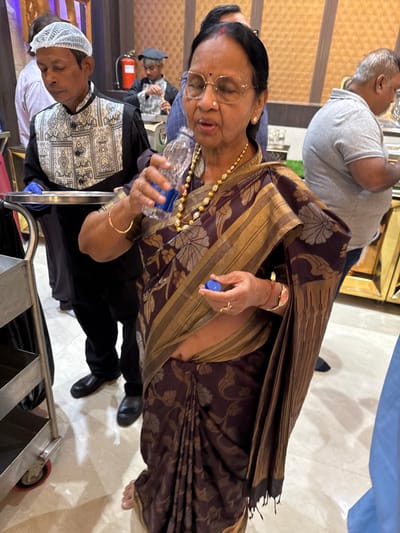 The image captures an indoor scene, likely a banquet hall or event space in New Town, India, characterized by patterned walls and light-colored tiled flooring.

The central subject is an elderly woman, wearing glasses, earrings, a gold necklace, a watch, and a dark brown saree with intricate gold/beige patterns and stripes. She holds a clear plastic water bottle with a blue cap, appearing to be drinking from it or preparing to do so. Her expression is neutral and focused on the bottle.

In the midground, to her lower left, a man in a black and white patterned chef's jacket or uniform, along with a white hairnet, stands near a metallic serving cart or trolley. His eyes appear closed or downcast. Another man in a grey polo shirt is partially visible in the background, facing away from the camera. Further in the background, a man wearing a cap is also partially obscured. A fire extinguisher is visible on the wall behind the man in the chef's uniform.

The overall activity suggests a social gathering or event, possibly catered, given the presence of the man in uniform and the serving equipment. The lighting appears artificial, indicating an indoor setting where the time of day is not evident. No visible text is present in the image.
