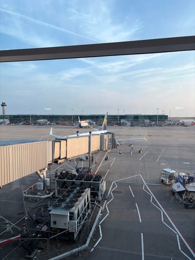 This image shows an aerial view of an airport tarmac, taken from inside a building, likely a terminal, looking out through a window. The scene is set on a sunny day with a clear blue sky dotted with wispy clouds. In the foreground, a jet bridge extends towards an airplane. The airplane has "RYANAIR" written on its tail fin. Several people can be seen on the tarmac, some walking near the airplane and others standing by vehicles.
Various airport vehicles and equipment are visible on the tarmac, including what appears to be a baggage tug or service vehicle with "BLUE HANDLING" printed on its side and the number "233". Further down the tarmac, more aircraft and airport infrastructure, including terminal buildings with large windows, are visible. A control tower can be seen in the distance on the left side of the image. The ground is paved with cobblestones in some areas and asphalt in others, marked with white lines indicating taxiways and parking spots. The overall atmosphere appears calm and functional, typical of an active airport environment. The time of day seems to be daytime, given the bright sunlight and clear sky. The location is identified as Uttlesford, United Kingdom.