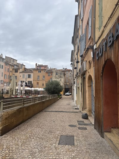 The image depicts a cobblestone street in Aix-en-Provence, France, under an overcast sky. The foreground features a narrow paved path containing several rectangular and circular utility access covers. On the right, a building displays a facade of vertical wooden panels with an arched entry, partially revealing the sign "CUTBA". A black plastic refuse bag rests at the base of this building. To the left, a concrete retaining wall topped with a metal railing borders a lower-level outdoor dining area. This area is furnished with multiple folded light-colored patio umbrellas, tables, and chairs, alongside a small green tree. No individuals are present within the scene. In the background, a dense cluster of multi-story buildings, characterized by varied pastel-colored facades and numerous shuttered windows, rises. A narrow street in the distance shows two white vehicles parked.