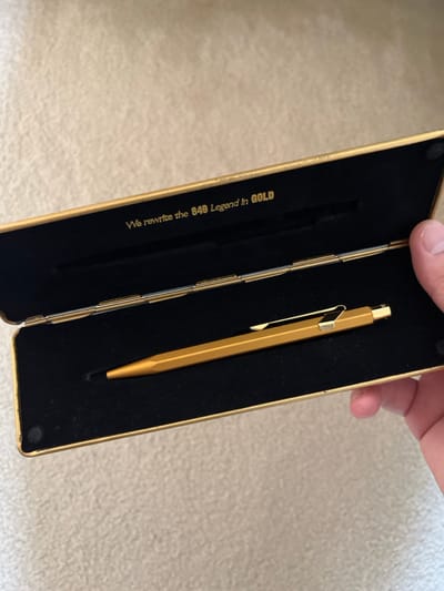 The image displays a close-up, slightly angled shot of an open, rectangular golden case holding a writing instrument. A human hand, with part of the thumb and fingers visible, is holding the case open from the top right.

The case itself has a metallic gold exterior and is lined with black velvet on the inside. It appears to be a presentation or gift box. On the right side of the open case, nestled securely in the black velvet, is a sleek, golden writing instrument, likely a pen or a mechanical pencil. It features a hexagonal or octagonal barrel shape and a matching golden pocket clip. The tip of the pen is visible at the bottom right.

On the left flap of the open case, gold-colored text is visible against the black velvet lining. The text reads: "We rewrite the 849 Legend in GOLD".

The background is a soft-focus, light-colored surface, possibly a carpet or a rug, suggesting an indoor setting. The lighting is even, highlighting the metallic sheen of the case and the pen. The overall impression is one of showcasing a premium, golden-themed writing tool.