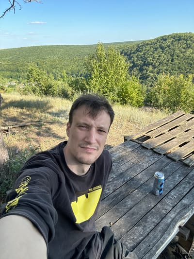 A man is taking a selfie while sitting on a wooden structure overlooking a vast, dense forest. The man is wearing a black long-sleeved shirt with a large yellow graphic design on the front, featuring the words "THE ORIGINAL" and a smiley face. He has short brown hair and a light beard.
The wooden structure appears to be made of weathered planks, possibly a table or a viewing platform. A partially consumed can of drink with blue and silver labeling rests on the wood near the man. The background reveals a sprawling expanse of green trees under a clear blue sky. In the distance, faint outlines of wind turbines are visible on the horizon, suggesting an open, elevated area.
The lighting indicates it is daytime, likely late morning or early afternoon, with the sun casting shadows on the wooden surface. The overall scene conveys a relaxed, outdoor atmosphere. The man's expression is neutral, and the composition suggests he is enjoying the view and the moment. The location is identified as Velykozalissia, Ukraine.