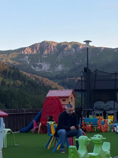 In Katun Bačko brdo, Montenegro, a man with a grey bun hairstyle sits on a blue plastic chair in a playground. He is wearing a black zip-up jacket and blue jeans, and is looking down at a mobile phone in his hands. The playground is equipped with a red plastic slide, various colorful children's chairs and tables, and several green dinosaur-shaped rocking toys. In the background, a trampoline structure with a black net is visible, along with a small, orange and red playhouse. A wooden fence encloses the playground area. Beyond the playground, a lush green forest covers the lower slopes of a rugged, rocky mountain, which is illuminated by the warm glow of the setting sun. The sky is clear and a pale blue.
