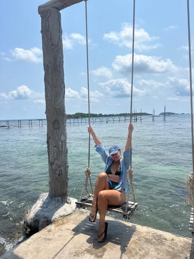 A single adult female is seated on a rustic wooden swing positioned over a concrete platform, adjacent to clear blue-green water, in Thành phố Phú Quốc, Vietnam. The female wears a dark green bikini, an unbuttoned light blue long-sleeved shirt, a blue baseball cap, black flip-flops, a light-colored choker necklace, and a pink wristband on her left wrist. Her legs are crossed at the ankles, and her arms are raised, gripping the swing ropes. She is smiling and looking towards the camera. The swing's ropes are suspended from a horizontal beam supported by a large, textured vertical post, appearing to be concrete sculpted to resemble wood, situated on the left side of the frame. The background features calm sea water under a bright blue sky with scattered white clouds. In the mid-distance, a long, rustic wooden pier-like structure extends across the water, and further right, a small forested island and additional vertical wooden poles rise from the sea.