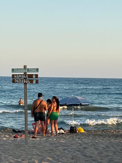 This media file depicts a summer day at the beach in Castelldefels, Spain. The scene is dominated by the ocean and a sandy beach, with a few people enjoying the seaside. The image is rotated, showing the sky on the left and the ocean on the right.

In the foreground, a wooden signpost stands on the sand. The sign has three wooden planks with text. The top plank, in green, reads "SUMMER TIME." The middle plank, in brown, reads "HAMACA" and the bottom plank, also in brown, reads "PARASOL."

Further back, near the water's edge, a group of people are visible. A man wearing dark shorts and a dark strap across his chest is looking towards the ocean. Beside him, a woman in a green skirt and bikini top is also present, along with another woman. Several beach items, including bags and a yellow object, are scattered on the sand.

A striped umbrella, partially visible, is set up on the beach. In the background, the ocean waves are gently rolling towards the shore, suggesting a calm day. The lighting indicates it is likely late afternoon or early evening, with the sun casting a soft glow. The overall mood is relaxed and summery, typical of a beach outing.