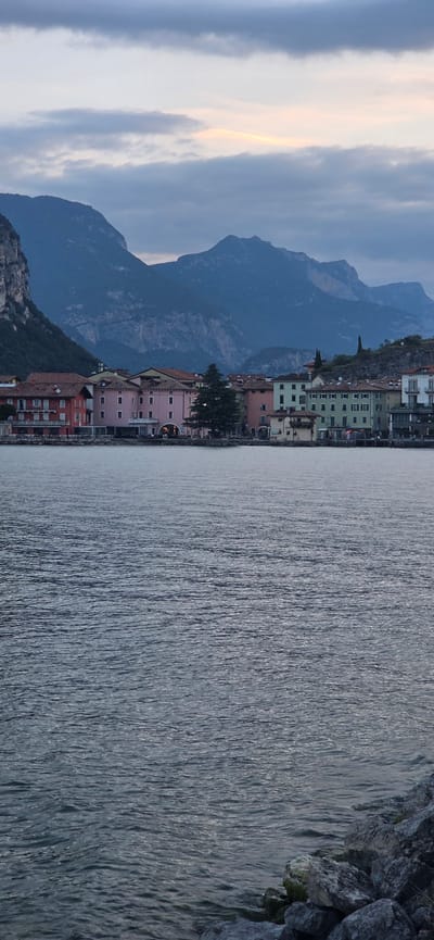 The image depicts a waterfront view in Torbole, Italy, with a calm body of water in the foreground and a town with mountains in the background. The water has small ripples and appears to be a lake or a bay. Along the shore, there is a line of buildings with colorful facades in shades of red, pink, green, and white. Some buildings have multiple stories and visible windows. Behind the town, a range of large, rugged mountains rises steeply, their peaks shrouded in a hazy, muted light. The sky is overcast with clouds, with a faint glow of sunrise or sunset visible on the horizon. The lower right corner of the image shows a rocky shoreline. There are no people or specific actions visible in the scene.