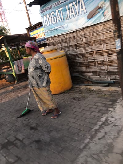 An elderly Indonesian woman with a pink and white headscarf and a gray floral patterned shirt is sweeping a wet, tiled walkway with a green-bristled broom. She is wearing a long yellow floral patterned skirt and blue flip-flops. To her right is a large, bright yellow water tank. Behind the woman and the water tank is a wall constructed of woven bamboo. A banner hangs above the bamboo wall, with blue text that reads "RESMI WEL JANGAT JAYA" and further text in a smaller font. The scene appears to be outdoors, likely in a public area in Glagah Arum, Indonesia, given the banner's mention of "Glagah harum Porong." The lighting suggests it is late afternoon or early morning.