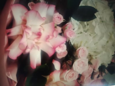 A close-up, slightly blurry photograph captures a bouquet of roses in various shades of pink and white. The flowers are densely packed, with some larger, open blooms in the foreground and smaller buds interspersed. Dark green leaves are visible in the background, adding contrast to the soft floral arrangement. The lighting appears to be ambient and diffused, creating a gentle, romantic atmosphere. The image does not provide any discernible details about the city of Qunduzak, Uzbekistan, or any specific location within it, nor does it show any people, actions, or interactions.