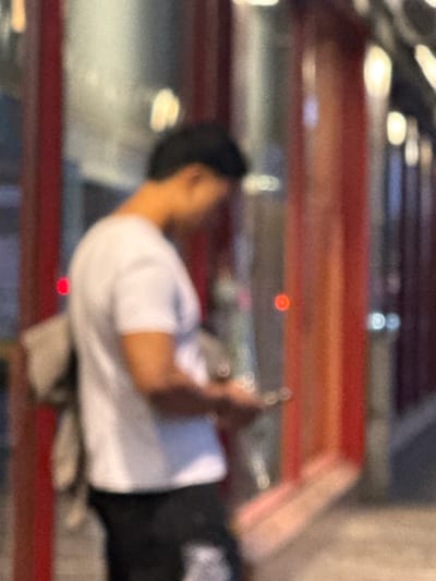 The image, taken at an unknown location in Sesto San Giovanni, Italy, shows a person in the foreground, partially obscured and out of focus. The individual is wearing a white t-shirt and dark pants, and appears to be holding a mobile device. The background features vertical red structures with indistinct lighting, suggesting an urban outdoor setting. The overall image quality is low with significant blur and grain.