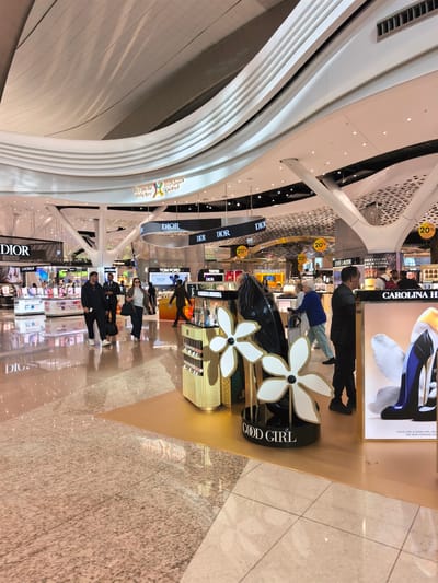 The image captures the interior of the Abu Dhabi Duty Free in Al Matar, United Arab Emirates. A polished, reflective floor indicates the high foot traffic and the upscale nature of the shopping area. The ceiling features an intricate, modern design with curved white panels and recessed lighting.
Several retail displays are visible, showcasing brands like Dior, Tom Ford, and Carolina Herrera. In the foreground, a large display promoting "Good Girl" perfume with large flower-shaped decorations is prominently featured. Various shoppers are present, browsing the displays and walking through the space. Some individuals are wearing masks, reflecting ongoing health precautions. The lighting is bright and even, creating a welcoming atmosphere for shoppers. The scene conveys a sense of bustling activity in a contemporary airport retail environment.