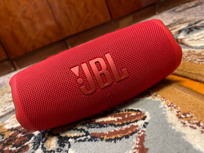 A close-up, angled shot shows a red JBL portable speaker resting on a carpet with a brown and tan patterned design. The speaker is cylindrical with a textured fabric exterior. The JBL logo is prominently displayed in a raised, metallic red on the side of the speaker. The background is blurred and appears to be a wooden surface. The overall scene suggests an indoor setting, possibly a home. There is no indication of the city of Bogdanovich, Russia, in the image.