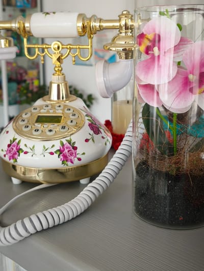 The image depicts a vintage-style rotary telephone adorned with pink roses and gold accents, placed on a grey surface. Adjacent to the telephone is a clear glass vase containing a floral arrangement of pink orchids and dried plant material, resting on black granules. The background is blurred but suggests an indoor setting with shelves holding various items. The scene does not provide any information about the city of Nevinnomyssk, Russia, or any specific actions or interactions.