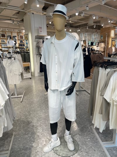 The image shows a male mannequin dressed in a white, loose-fitting linen outfit, consisting of a short-sleeved button-up shirt and shorts. The mannequin is wearing a white fedora hat, white socks, and white sneakers. It is standing on a circular glass platform in a retail clothing store. The store appears to be well-lit with overhead lights and features racks of clothing in neutral tones. The floor is covered in a hexagonal tile pattern. In the background, other mannequins and clothing displays are visible, suggesting this is a men's fashion section. There are no people visible in the image. Based on the context provided, this scene is located in Marcianise, Italy, within a retail store environment.