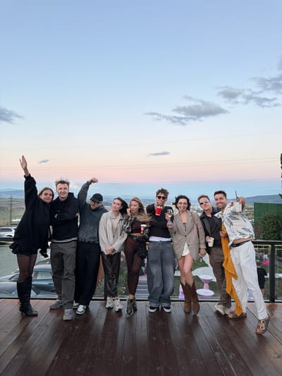 A group of ten young adults are posing for a photo on a wooden deck, with a hazy, sunset-colored sky and distant hills in the background. The people are dressed in casual, trendy attire, and several are holding drinks. The location is identified as Lisi, Georgia, likely an outdoor event space. The scene suggests a relaxed, social gathering.