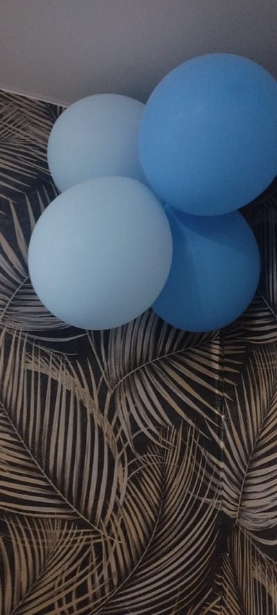 The image shows a close-up view of four light blue and blue balloons against a background of dark wallpaper with a palm leaf pattern. The balloons are clustered together, with some overlapping. The lighting is dim, and the image quality is moderate, suggesting it was taken indoors. There is no indication of the city of Покосное, Russia, or any other specific location. The scene appears to be staged, likely for a celebration.