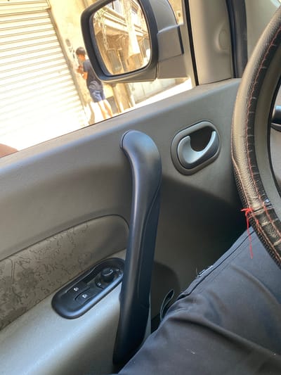 The image depicts the interior of a car in Baraki, Algeria, focusing on the driver's side door panel. The door panel is light gray and features a door handle, a power window switch, and a lock switch. A steering wheel with a black leather cover and red stitching is visible at the top of the frame. A person wearing black pants is partially visible next to the steering wheel. The side mirror reflects a scene outside the car: a person wearing shorts and a t-shirt is lying on the ground in front of a building with a corrugated metal facade.