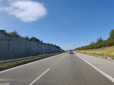 The image shows a bright, sunny day on a highway in Golemo Buchino, Bulgaria. The perspective is from inside a vehicle traveling on the road. To the left, there is a concrete retaining wall that slopes upwards, topped with green trees and vegetation. A metal guardrail runs along the edge of the road in front of the wall. The highway itself is paved with asphalt and has white lane markings. In the distance, other vehicles can be seen, including a dark truck, a white car, and a silver SUV. The right side of the highway features a grassy embankment covered with dry yellow grass and more green trees. The sky is a clear, vibrant blue with a large, fluffy white cloud in the upper left quadrant. The overall scene depicts a typical highway environment in a rural or semi-rural setting.