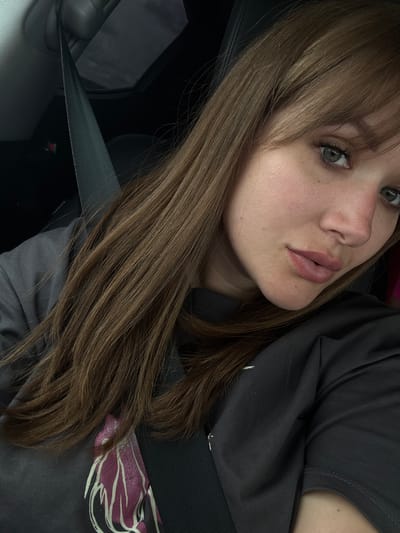 A selfie-style close-up image shows a young woman with shoulder-length brown hair and fair skin, wearing a gray t-shirt and a seatbelt. Her face is slightly angled towards the camera, highlighting her green eyes, freckles, and full lips. The background is dark and indistinct, but shows elements of a car interior. There is no verifiable information that indicates this photo was taken in the city of Troitskoe, Russia.