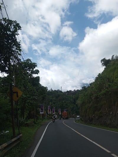 The image displays a two-lane asphalt road curving through a mountainous, heavily vegetated area in Gubbisamamidi, India. The sky is partly cloudy with blue patches and scattered white cumulus clouds.
On the left side of the road, dense green foliage, trees, and bushes are visible. A yellow diamond-shaped road sign with a black left-turn arrow is affixed to a pole, alongside which numerous utility wires are entangled. A rusted metal guardrail runs along the road's edge. Further along the left side, at least four vertical purple banners with white text/graphics are mounted on poles. A white SUV and a person riding a motorcycle are observed on the road, moving away from the camera.
The right side of the road is bordered by a steep, lush green hillside, featuring dense vegetation and some exposed rock. In the mid-ground, a large tan-colored truck, possibly a flatbed carrying a brown load, is visible heading away from the viewer. Other smaller vehicles are present further down the road.
The background consists of forested hills and mountains, partially obscured by clouds. A structure resembling a telecommunications tower is discernible on a distant peak. The road itself has a solid white line on its left edge and a solid yellow line marking the center.