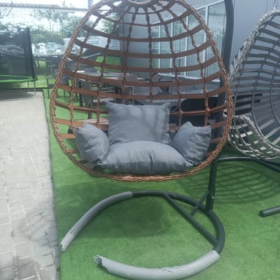 In Briceni, Moldova, an outdoor seating area features a brown wicker egg-shaped hanging chair with grey cushions, supported by a black metal stand. The stand's base is partially covered in grey foam padding, some of which appears damaged and torn. The chair is situated on artificial green turf, bordering a paved walkway made of light grey rectangular stones. In the background, other rattan-style chairs and tables are visible, along with a building with large windows. To the right of the brown chair, another grey wicker hanging chair is partially visible. A black trampoline with a green mat is also present in the background, to the left of the main chair. The scene suggests a commercial or recreational outdoor space.