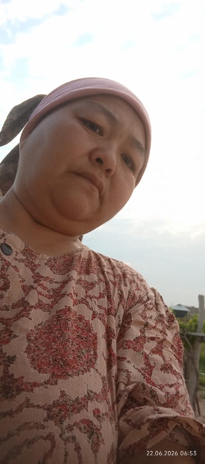 A medium-shot selfie captures a person with a light complexion from the chest up, looking down towards the camera. They are wearing a light pink headscarf and a long-sleeved garment with a red and beige floral pattern. The background is a bright, hazy sky with subtle cloud formations, suggesting an outdoor setting. A wooden post and a hint of a small building can be seen to the right. The lower right corner displays a timestamp: "22.06.2026 06:53". The image was taken in or near Chon-Kara, Kyrgyzstan.