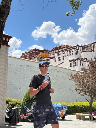 This outdoor image captures a bright, sunny daytime scene with a clear blue sky dotted with fluffy white clouds. The main subject is a man standing in the foreground, positioned slightly to the right of the frame. He is wearing a black t-shirt, patterned black and grey shorts, a blue baseball cap, and sunglasses, and he holds a patterned water bottle in his right hand. He has a slight smile on his face. Colorful bracelets and a watch are visible on his left wrist.
Behind the man, a light-colored wall runs horizontally across the mid-ground. Beyond the wall, a strip of vibrant green bushes and a tree with brown, autumn-like leaves are visible. To the left, a large, majestic building with distinct Tibetan architecture dominates the background. This multi-storied structure is predominantly white with red and dark brown sections, featuring many small, dark windows. Its appearance strongly suggests it is the Potala Palace in Lhasa, consistent with the Chengguan District, China location. Another tree with green foliage is visible in the upper left corner of the sky.
On the paved ground near the man's feet, to his right, are several items including a blue and white bag or piece of luggage, on which Chinese characters are visible, including "藏" (Zàng), a common character for Tibet. A black bag is also present nearby. Further to the right, a wooden bench is visible. The overall scene depicts a tourist enjoying a sunny day at a historic landmark.