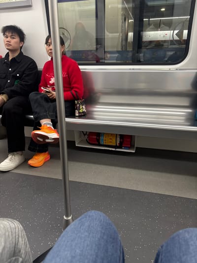 Two people are seated on public transportation. One wears a red sweater and orange sneakers, while the other wears a black jacket and white sneakers. A fire extinguisher is visible on the wall behind them. A metal handrail extends across the scene. The lower portions of two more people, wearing jeans, are also visible. The interior of the transit car includes a window and metallic paneling.