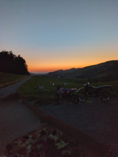 At dusk, two dirt bikes are parked on a gravel road in a rural area of St. Peter, Germany. The sky displays a gradient of blue to orange hues as the sun sets behind rolling hills covered in trees and greenery. In the foreground, a partially visible pizza box rests on the road. A paved path winds through a grassy field to the left of the motorcycles, bordered by trees. The lighting suggests late evening, with a clear sky.