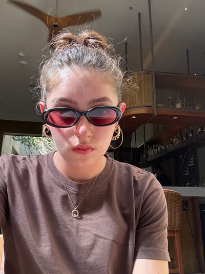 A person with their hair pulled back, wearing black-framed sunglasses with red lenses, faces the camera. They wear a brown t-shirt and a delicate necklace with a circular pendant. Large, gold hoop earrings adorn their ears, and a small nose piercing is visible. The background suggests an indoor setting with a bar or restaurant area, a ceiling fan, and natural light filtering through a window.